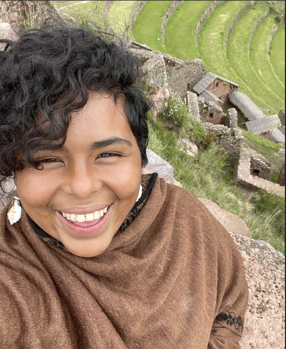 A woman smiling joyfully, with short black/brown hair, a brown shawl, and seashell earrings.