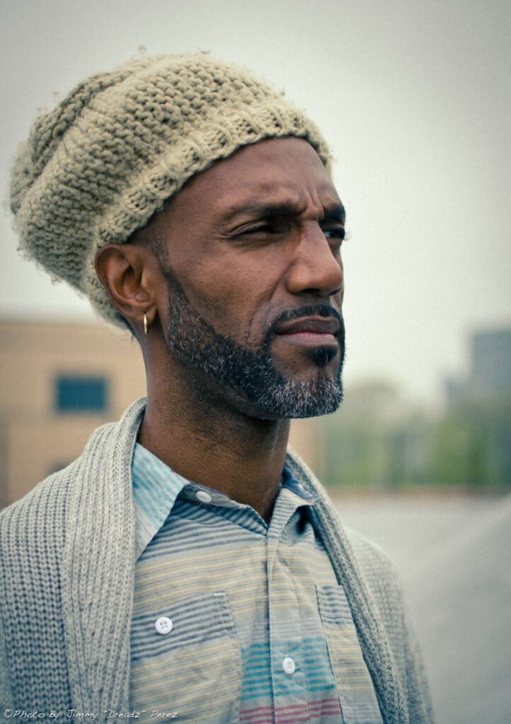 A photo of a black man wearing a head covering, grey shirt, and gazing out into the distance.